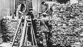 Fotografía en blanco y negro que muestra a cinco hombres trabajando en la construcción de un muro de ladrillos, con andamios de madera a la izquierda