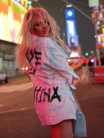 Una mujer rubia sonríe y mira hacia atrás, viste una camisa blanca con manchas celestes y letras negras. Se encuentra en una calle iluminada por luces de neón