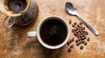 Una jarra de café con filtro, una taza de café negro con gotas de aceite, granos de café tostados y una cuchara metálica sobre una mesa de madera.