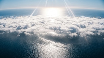 Vista aérea del océano Pacífico con una capa de nubes blancas sobre el agua, un cielo azul y los intensos reflejos del sol en la superficie marina y las nubes.
