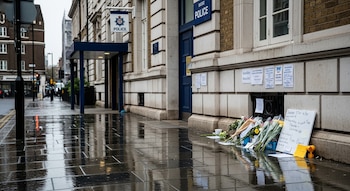 Fachada de comisaría de policía, cartel con el logo de "Police", puerta azul, acera mojada, flores, ramos, cartel blanco, notas en la pared, edificios, calle.