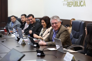 Grupo de personas en una mesa de conferencias. Un hombre de gafas sonríe al frente. La mesa tiene laptops, tabletas y micrófonos. Se lee 'DE LA REPÚBLICA'