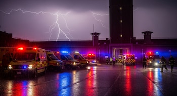 Múltiples ambulancias y vehículos de emergencia con luces encendidas estacionados frente a una prisión durante una tormenta con rayos, pavimento mojado y personal de rescate.