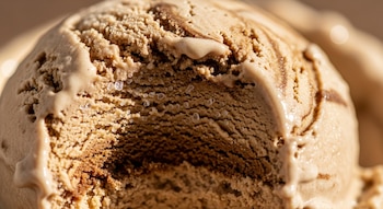 Detalle de la superficie de un helado de café y leche de almendras, mostrando su textura granulada y cremosa con vetas más oscuras y pequeñas gotas de agua derretida.
