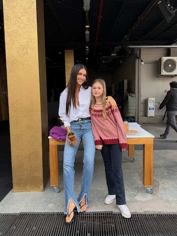 Zaira Nara, con camisa blanca y jeans, sonríe junto a una niña vestida con un suéter rosa y pantalón oscuro, de pie al aire libre