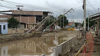 Postes de concreto inclinados sobre una zanja con agua y cables caídos frente a casas en una calle de tierra