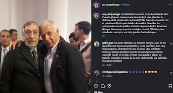 Dos hombres mayores, ambos con traje formal, posan sonrientes. Uno con gafas y barba, el otro con el brazo sobre su hombro. Al fondo, otras personas difuminadas