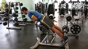 Un hombre vestido con camiseta azul y pantalones cortos grises realiza extensiones de espalda en una máquina en el interior de un gimnasio.
