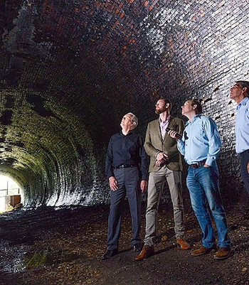 Cuatro hombres observan el techo de un túnel de ladrillos. Uno ilumina la estructura con una linterna. El suelo es oscuro y hay una apertura luminosa al fondo