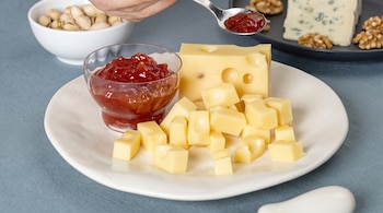 Mano con cuchara sirviendo confitura roja, un bol de confitura, un bloque de queso amarillo, cubos de queso, un plato con queso azul y nueces, un bol de pistachos.