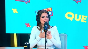 Mujer con auriculares negros y micrófono, con lágrimas en los ojos y mano derecha en el pecho, sentada frente a una taza en una mesa, con fondo azul brillante.