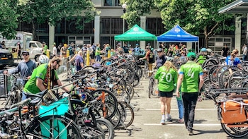 Un gran número de bicicletas, road bikes y hybrid bikes, estacionadas en un aparcamiento al aire libre con muchas personas y dos carpas en un día soleado