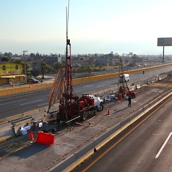 Vista aérea de maquinaria de perforación, un camión y barreras de concreto en el carril de una autopista. Un trabajador se encuentra cerca del equipo