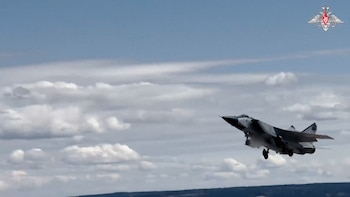 Un avión de combate gris, con el tren de aterrizaje extendido, asciende hacia un cielo azul con nubes blancas. En la esquina superior derecha, un emblema rojo y blanco