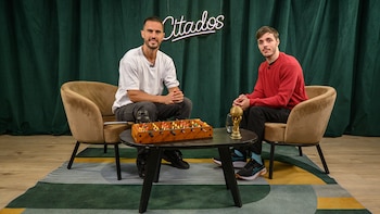 Dos hombres sentados en sillas club marrones, sonriendo. Entre ellos, una mesa de café con un futbolín, una copa y un vaso. Fondo de telón verde con rótulo 'Citados'