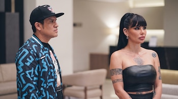 Giancarlo Cossío con gorra negra y chaqueta estampada junto a Angie Jibaja con top negro, ambos de pie mirando a la derecha en un interior.