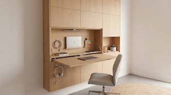 Un mueble de madera clara empotrado en la pared con un escritorio plegable abierto. Contiene un portátil, auriculares, suministros de oficina y una silla.