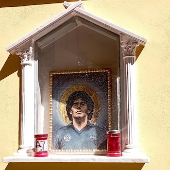 Pequeño santuario de pared con un retrato enmarcado de Diego Maradona en camiseta del Napoli, enmarcado con un halo dorado, y dos velas rojas
