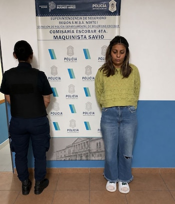 Una mujer con suéter verde y jeans, y un oficial de policía de espaldas, parados frente a un banner de la Comisaría Escobar 4ta. Maquinista Savio
