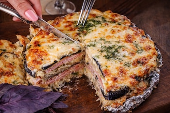 Una mano corta un pastel gratinado de berenjena, jamón y queso, envuelto en papel de aluminio, sobre una tabla de madera, con hierbas y hojas moradas.