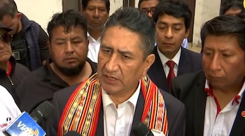 Primer plano de un hombre hablando a dos micrófonos. Lleva una camisa blanca, un saco oscuro y una bufanda de tela con diseño andino. Detrás hay otros hombres.