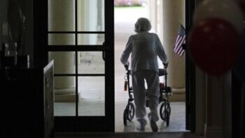 Uma bandeira americana e balões podem ser vistos no fundo de um idoso de cabelos brancos usando um andador para atravessar uma porta