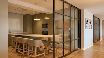 Cocina moderna con isla, taburetes beige, gabinetes verdes y electrodomésticos empotrados. Una partición de vidrio y metal divide el espacio, mostrando un comedor al fondo.