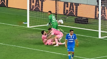 Dos futbolistas con uniforme rosa celebran en el césped cerca del marco mientras un jugador en uniforme azul camina en primer plano.