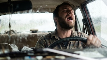 Un hombre barbudo con camisa a cuadros canta en un vehículo. Tiene la boca abierta y las manos en el volante. Las ventanas tienen gotas de lluvia.