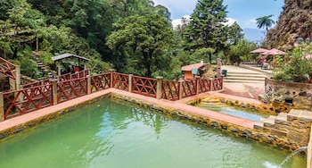 Piscinas termales de agua verde con barandas rojas. Rodeadas de senderos de piedra, árboles frondosos, vegetación y pequeñas estructuras con sombrillas