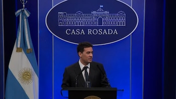 Un hombre con traje oscuro y corbata en un atril frente a una bandera de Argentina y un cartel con el dibujo de la Casa Rosada y su nombre