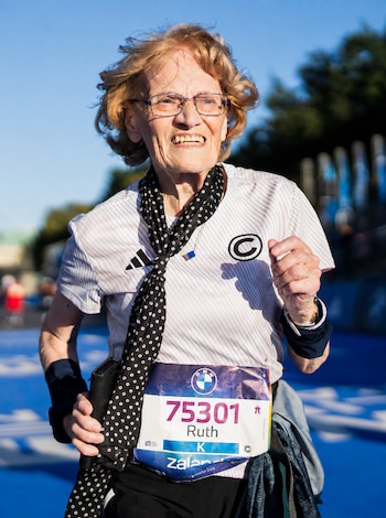 Una mujer mayor con anteojos, pañuelo a lunares y un dorsal con el número 75301 y el nombre Ruth corre en una pista azul