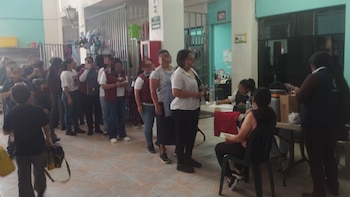 Larga fila de personas, incluidos estudiantes, esperando en un pasillo. Al final de la fila, personal médico en una mesa registra y vacuna a los asistentes