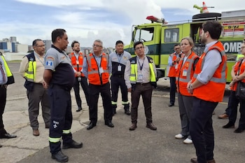 Un grupo de diez personas, algunas con uniformes y otras con chalecos naranja o amarillo, de pie en una pista de asfalto junto a un camión de bomberos