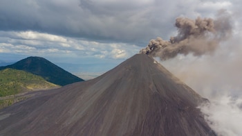 Vista aérea de un volcán gris oscuro en erupción con una columna de humo, junto a una montaña verde y un cielo nublado