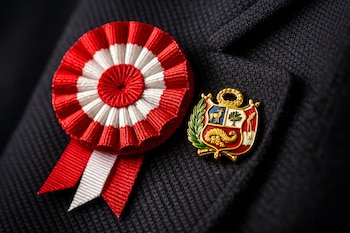 Escarapela de tela roja y blanca con franjas plisadas junto a un pin metálico del escudo nacional del Perú, prendidos en un saco oscuro.