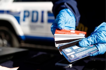 Primer plano de una mano enguantada de azul sosteniendo varias tarjetas de crédito frente a un vehículo de la Policía de Investigaciones (PDI) de Chile