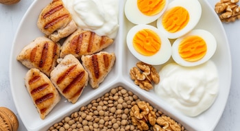 Plato blanco dividido con trozos de pollo a la plancha, huevos duros partidos, yogur griego, lentejas cocidas y nueces enteras.