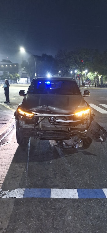 Automóvil Volkswagen oscuro con daños frontales graves en una calle por la noche, con luces de emergencia azules, faros encendidos y un oficial en el fondo