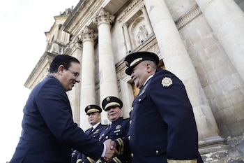Un hombre con traje oscuro y gafas estrecha la mano de un oficial con uniforme azul marino; dos oficiales más detrás y un edificio clásico con columnas al fondo