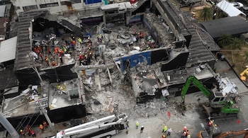 Vista aérea de un edificio parcialmente colapsado, mostrando escombros y numerosos rescatistas con cascos y chalecos de seguridad naranjas y amarillos. Un excavador verde y un camión elevador blanco son visibles.