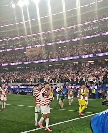 Gio Reyna, vistiendo uniforme blanco y rojo, envía un beso al público en un estadio de fútbol abarrotado con aficionados animando en las gradas
