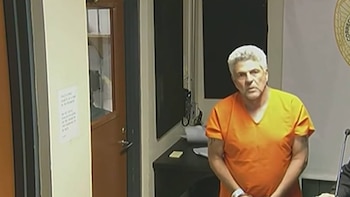 Hombre de cabello gris con uniforme naranja parado en una sala. Se ve un escritorio oscuro, una puerta de madera con ventana y un sello parcial en la pared