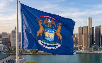 Primer plano de la bandera de Michigan azul con su escudo de armas, ondeando frente a un horizonte urbano con múltiples edificios altos junto a un cuerpo de agua