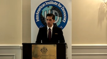 Hombre de traje y corbata detrás de un podio con placa Trump National Doral Miami. Fondo con estandarte del InterAmerican Institute for Democracy