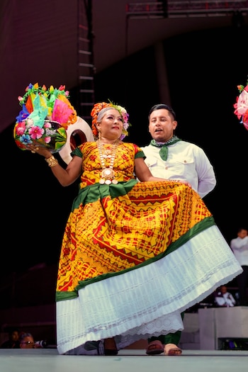 Mujer con vestido folclórico amarillo, rojo y verde, joyas, flores en el cabello y un recipiente decorado. Hombre con camisa blanca y pañuelo verde