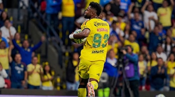 Un futbolista de espaldas con uniforme amarillo lleva un balón bajo el brazo ante la tribuna del estadio.