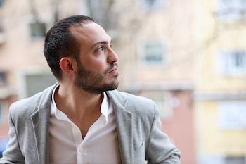 Primer plano de un hombre con barba corta y cabello oscuro, vestido con una chaqueta gris y camisa blanca, mirando hacia arriba y a la derecha