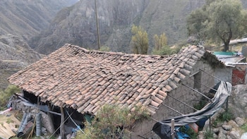 Casa rural con techo de tejas irregulares, paredes de barro y soportes de madera. En el fondo, un paisaje montañoso con árboles y vegetación