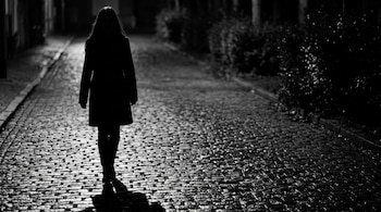 Silueta de una persona con cabello largo caminando sobre calle de adoquines mojados en la oscuridad. Imagen en blanco y negro.